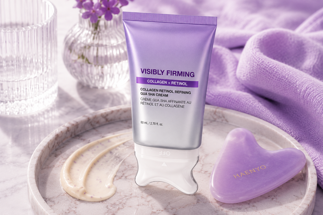 Haenyo Visibly Firming gua sha cream tube standing on a marble tray beside two swatches of the cream and a lilac heart-shaped gua sha stone, with violets in a fluted glass vase.