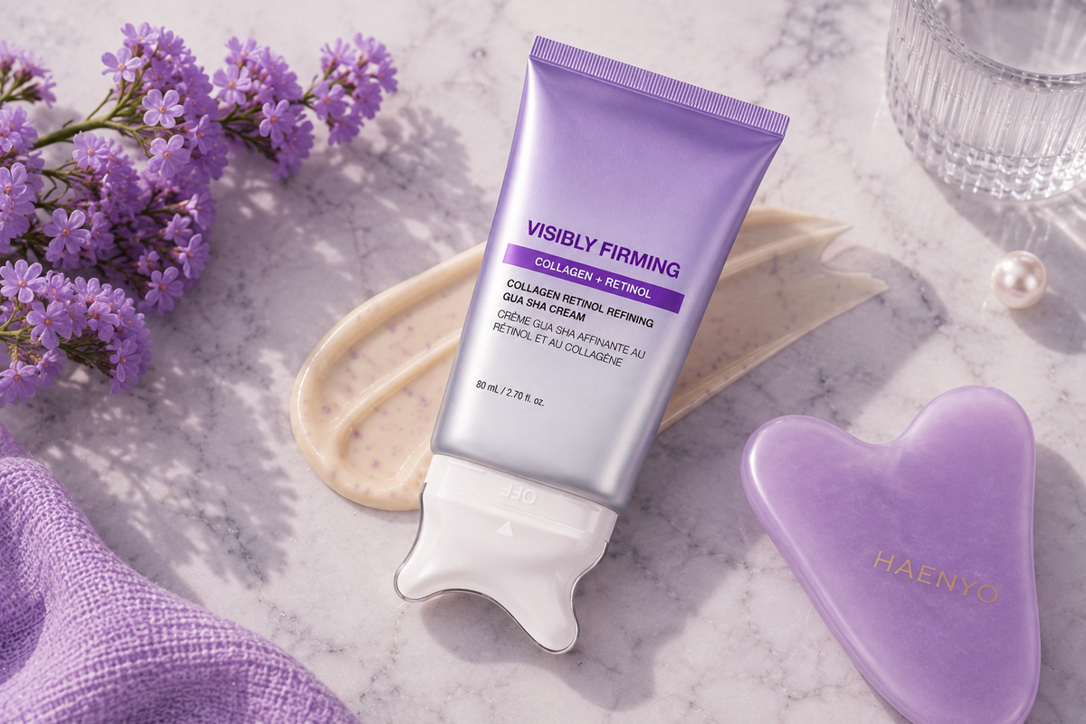 Overhead flat lay of the Haenyo cream tube on marble with a broad swatch of cream, a lilac heart-shaped gua sha stone, lilac blossoms and a cut-glass tumbler.